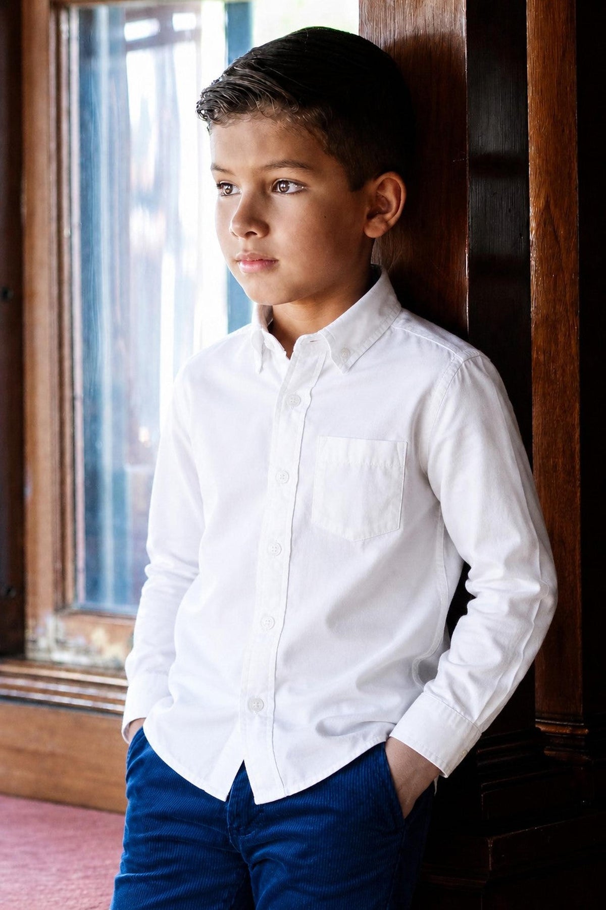 A boy stands by a window, wearing a classic white organic cotton stretch poplin shirt with a button-down collar and a chest pocket.