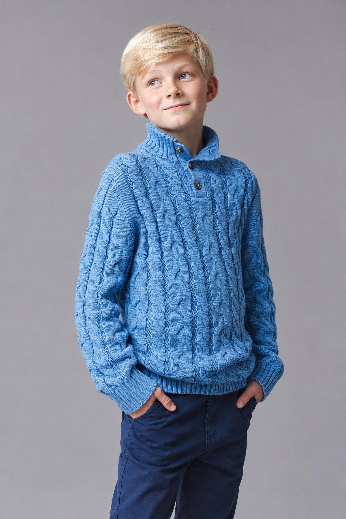 A boy models a medium blue organic cotton mock neck cable sweater with a button placket, featuring a textured knit design.