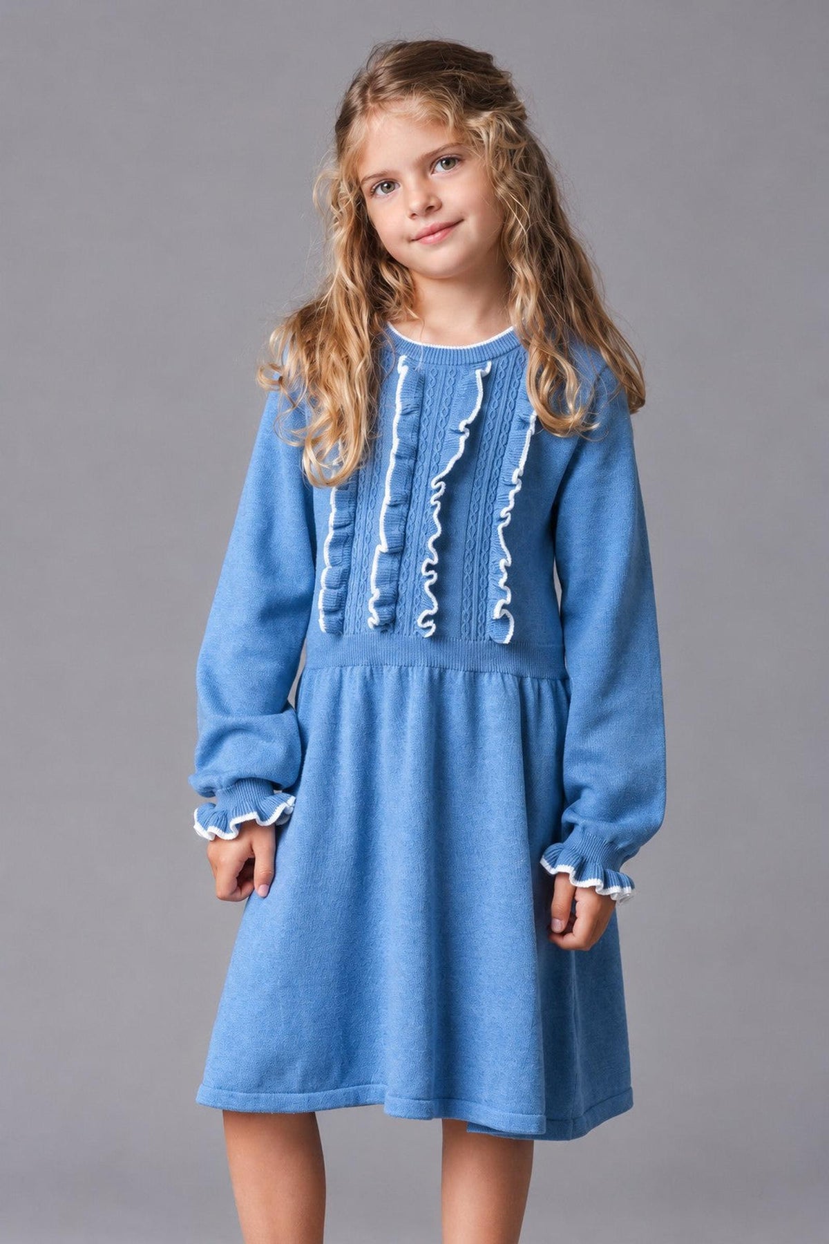 A young girl models a medium blue organic cotton sweater dress featuring a ruffled bodice and cuffs. The dress has a relaxed fit, perfect for comfortable wear in cooler weather.