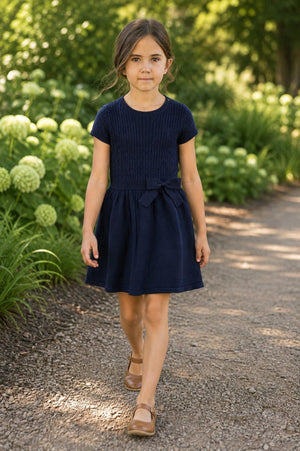 A girl walks on a gravel path surrounded by greenery, wearing a navy short sleeve cable knit dress with a bow at the waist.