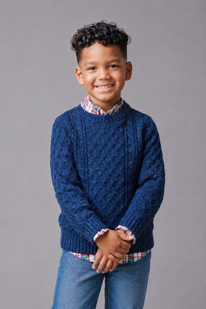 A young boy smiles while wearing a navy organic cotton cable knit fisherman sweater over a plaid shirt, paired with blue jeans, set against a neutral gray background.