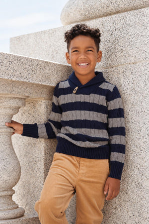A young boy smiles while leaning against a stone railing, wearing a navy and gray striped cable knit sweater.