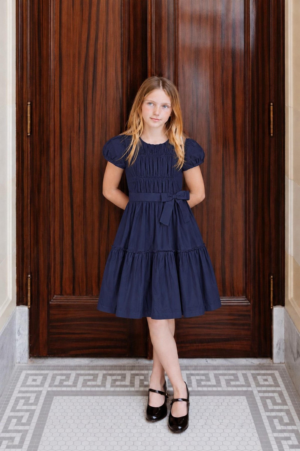 A young girl stands confidently in a navy organic cotton tiered dress with puff sleeves and a waist sash. The elegant setting features dark wooden doors and a patterned floor.