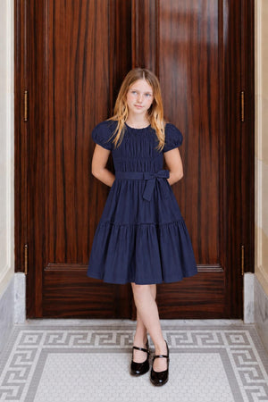 A young girl stands confidently in a navy organic cotton tiered dress with puff sleeves and a waist sash. The elegant setting features dark wooden doors and a patterned floor.
