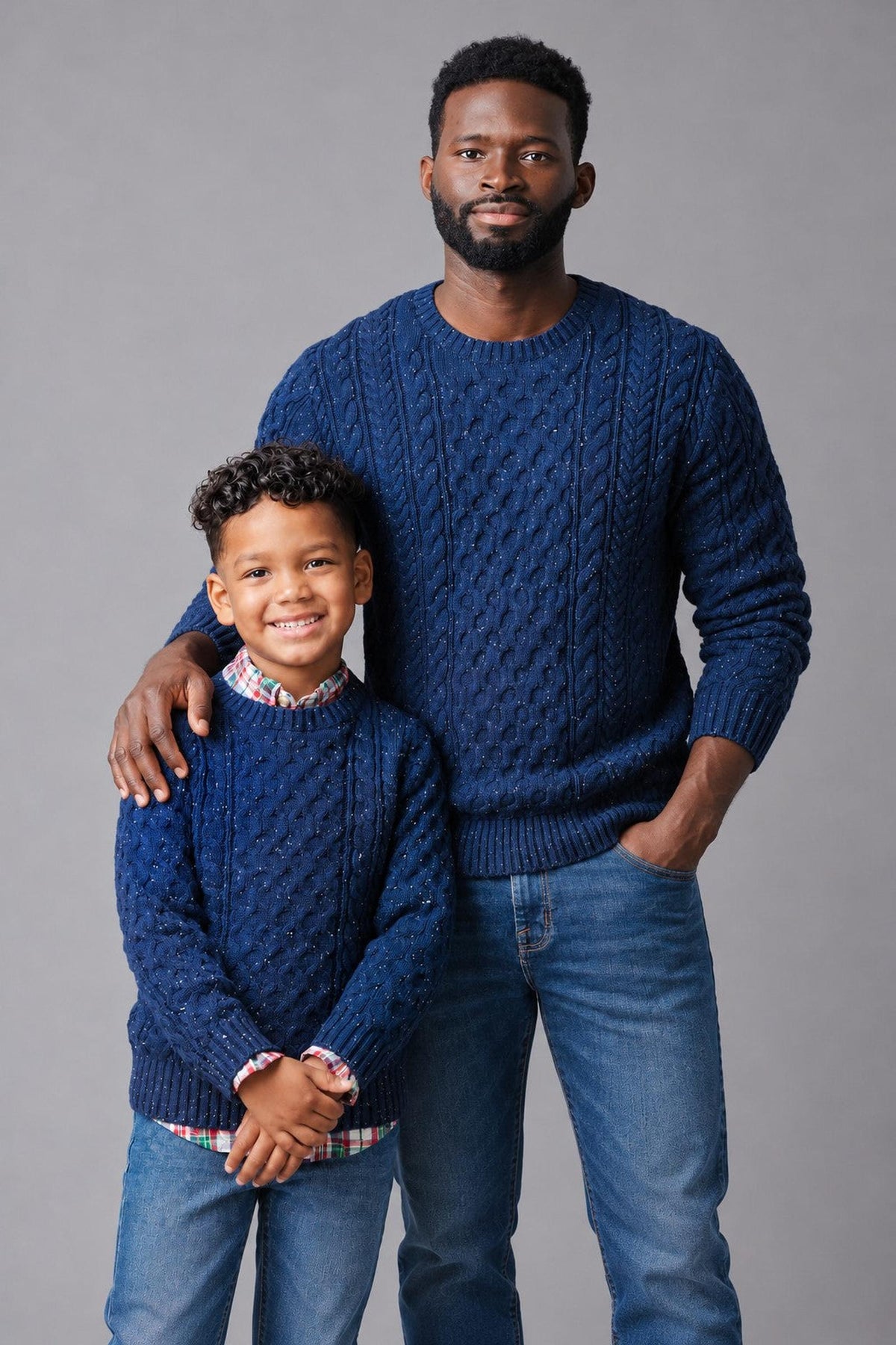 A man and a boy wear matching navy organic cotton fisherman sweaters, featuring allover cable designs. The setting has a neutral background, highlighting their coordinated outfits and family connection.