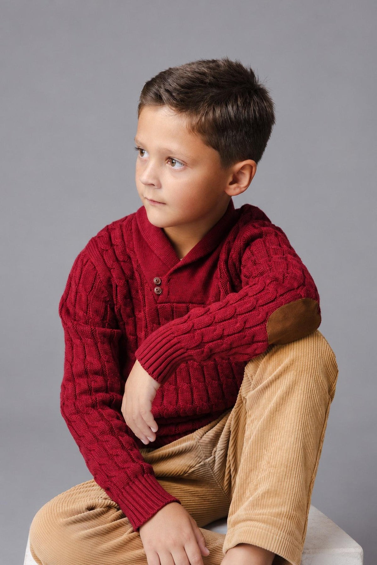 A young boy sits thoughtfully, wearing a deep oxblood organic cotton shawl collar cable sweater. The sweater features a button closure and elbow patches, paired with light corduroy pants.