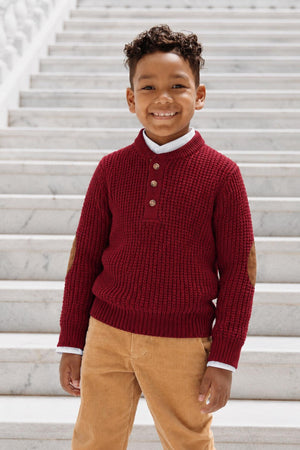 A smiling young boy wears a cozy, oxblood waffle-knit sweater henley featuring faux suede elbow patches. He stands on marble steps, showcasing a stylish and comfortable look for children.