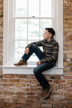 A boy sits on a windowsill, wearing a navy plaid organic cotton flannel shirt paired with dark jeans and brown shoes, against a backdrop of rustic brick walls.