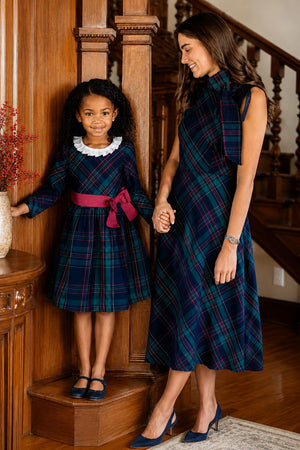 A woman and a girl stand together, both wearing elegant Scottish plaid dresses. The womans dress features a bow neckline.