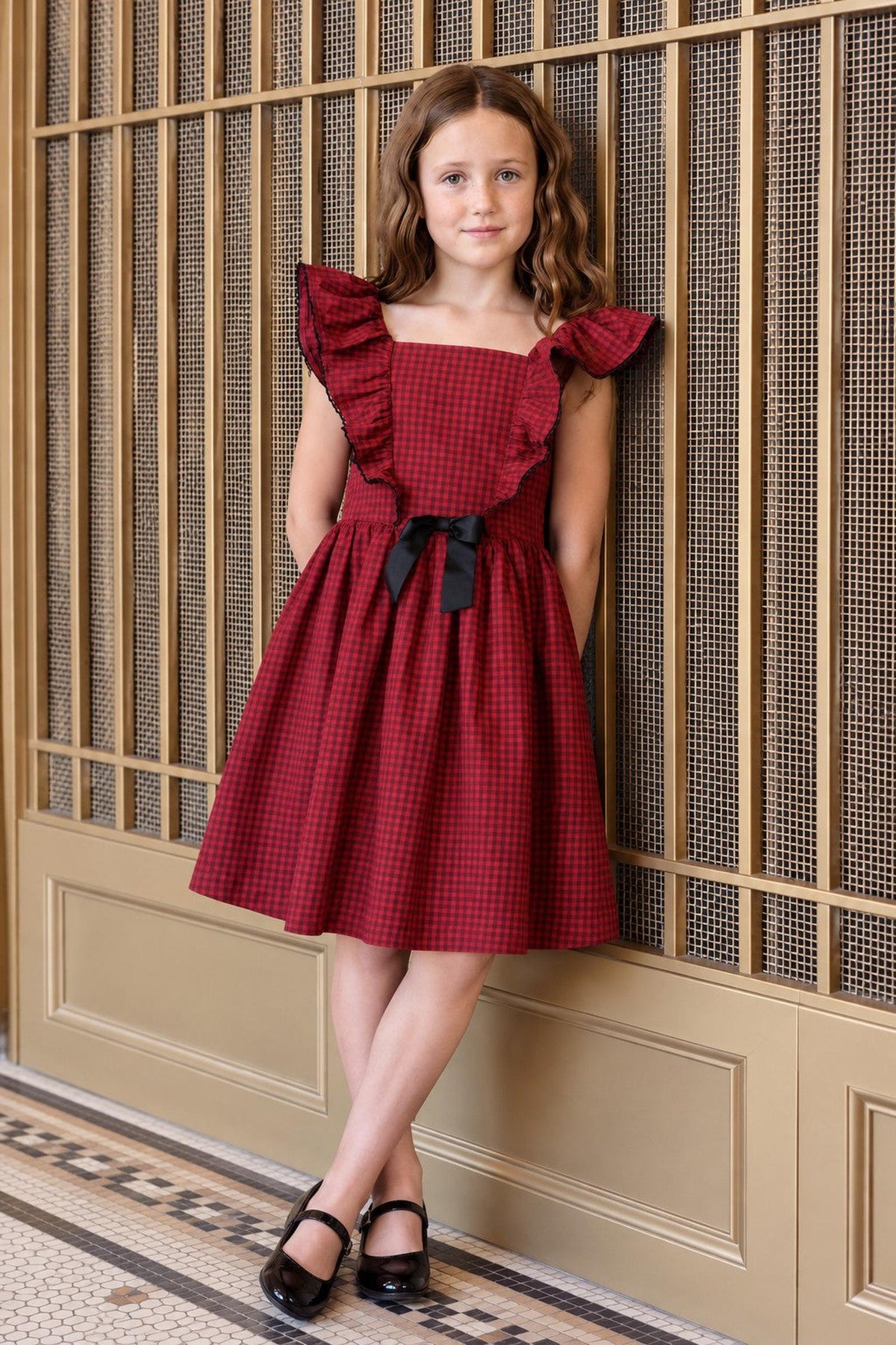 A young girl poses in a red gingham ruffle jumper party dress with a black bow, featuring ruffled sleeves and a flared skirt, against a decorative gold backdrop.