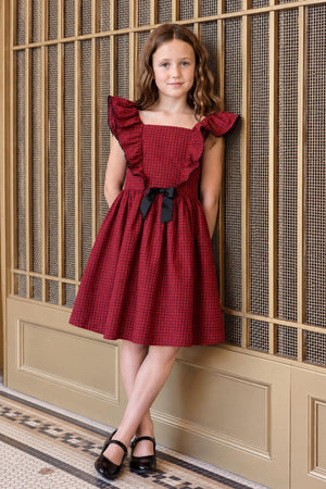 A young girl poses in a red gingham ruffle jumper party dress with a black bow, featuring ruffled sleeves and a flared skirt, against a decorative gold backdrop.