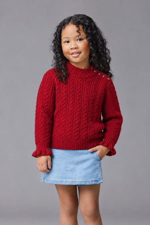 A young girl with curly hair smiles while wearing a cozy red cable knit turtleneck sweater with ruffle cuffs and gold button details, paired with a denim skirt against a.