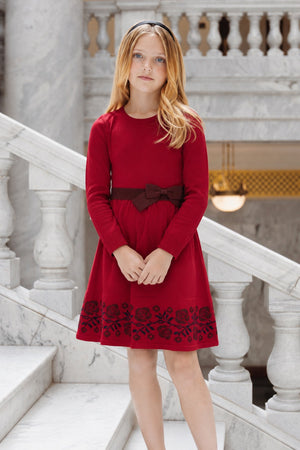 A young girl stands on a marble staircase, wearing a red long-sleeve skater dress with a decorative bow at the waist. The dress features floral embroidery along the hem.