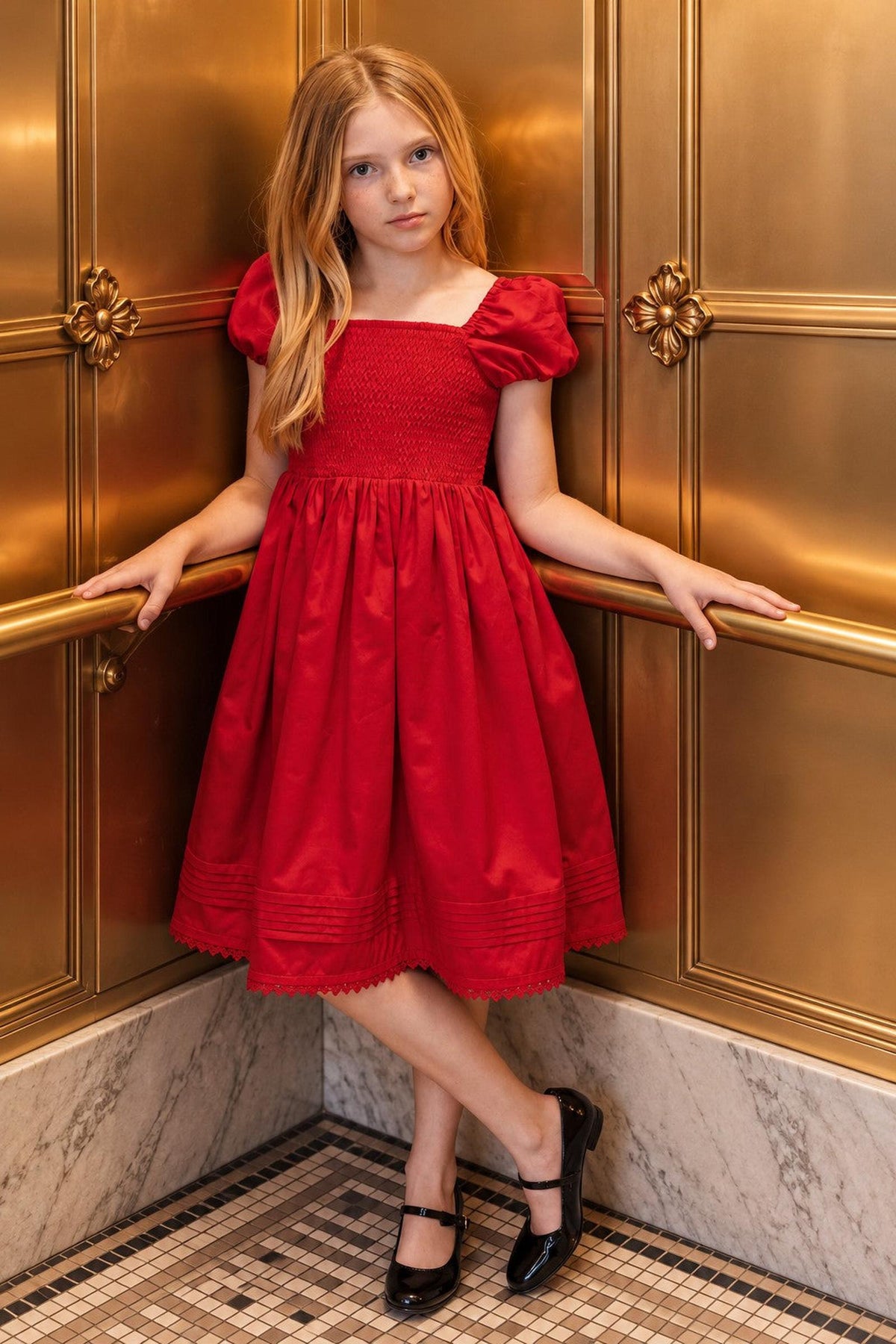 A young girl stands elegantly in a red organic cotton sateen dress with puffed sleeves and a pintucked hem, posing against a luxurious golden backdrop. She wears classic black shoes.