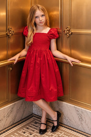 A young girl stands elegantly in a red organic cotton sateen dress with puffed sleeves and a pintucked hem, posing against a luxurious golden backdrop. She wears classic black shoes.
