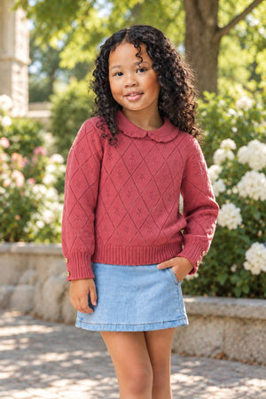 A young girl smiles while wearing a rosewood organic cotton sweater with a peter pan collar and diamond pattern, paired with a denim skirt, set against a blooming garden backdrop.