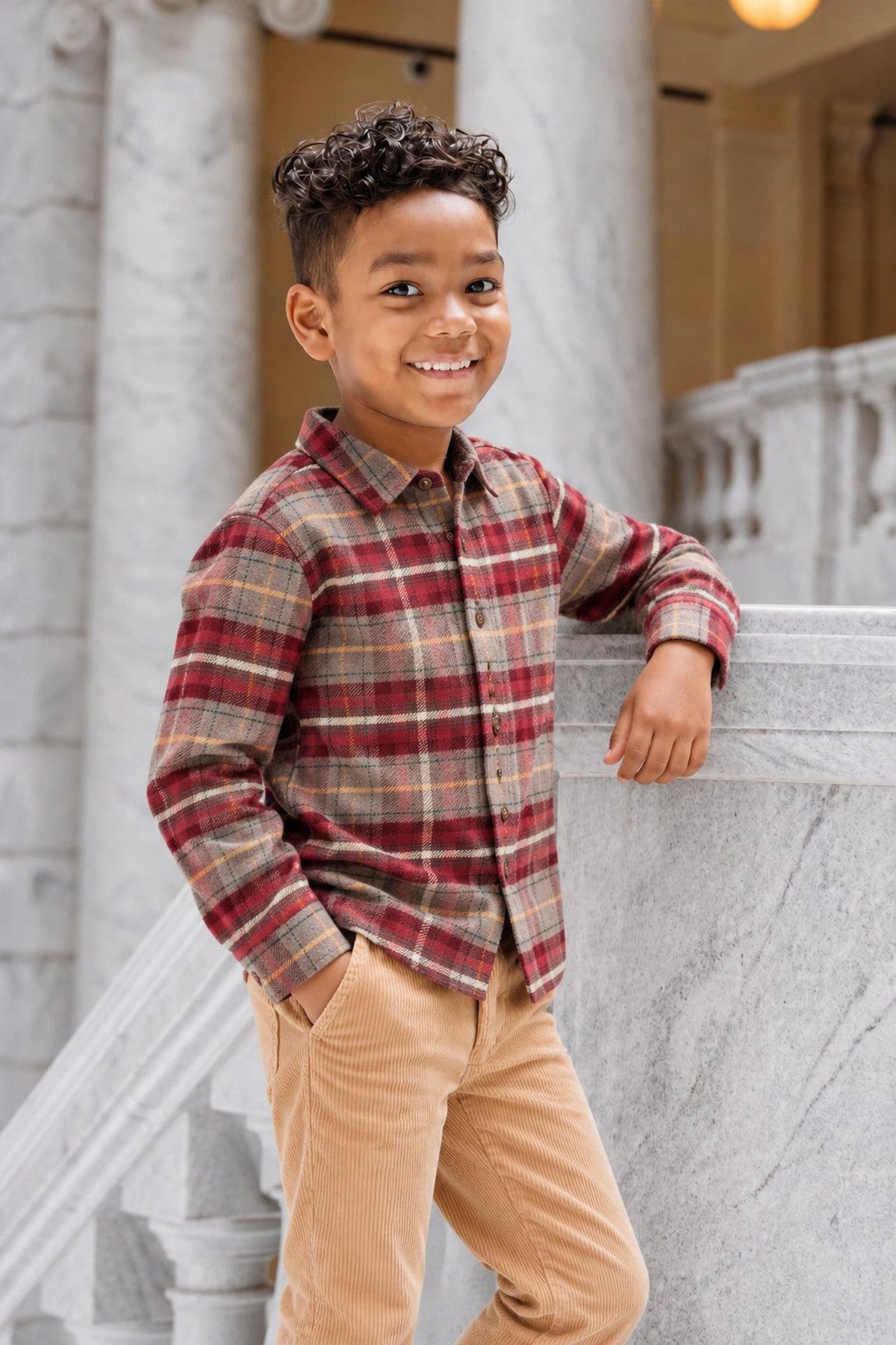 A young boy smiles while leaning against a marble staircase, wearing a red and beige plaid organic cotton flannel shirt.