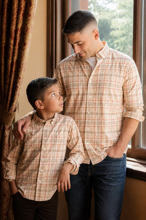 A father and son stand together by a window, both wearing matching plaid shirts in soft, organic cotton. The warm tones of the shirts complement the cozy interior setting.