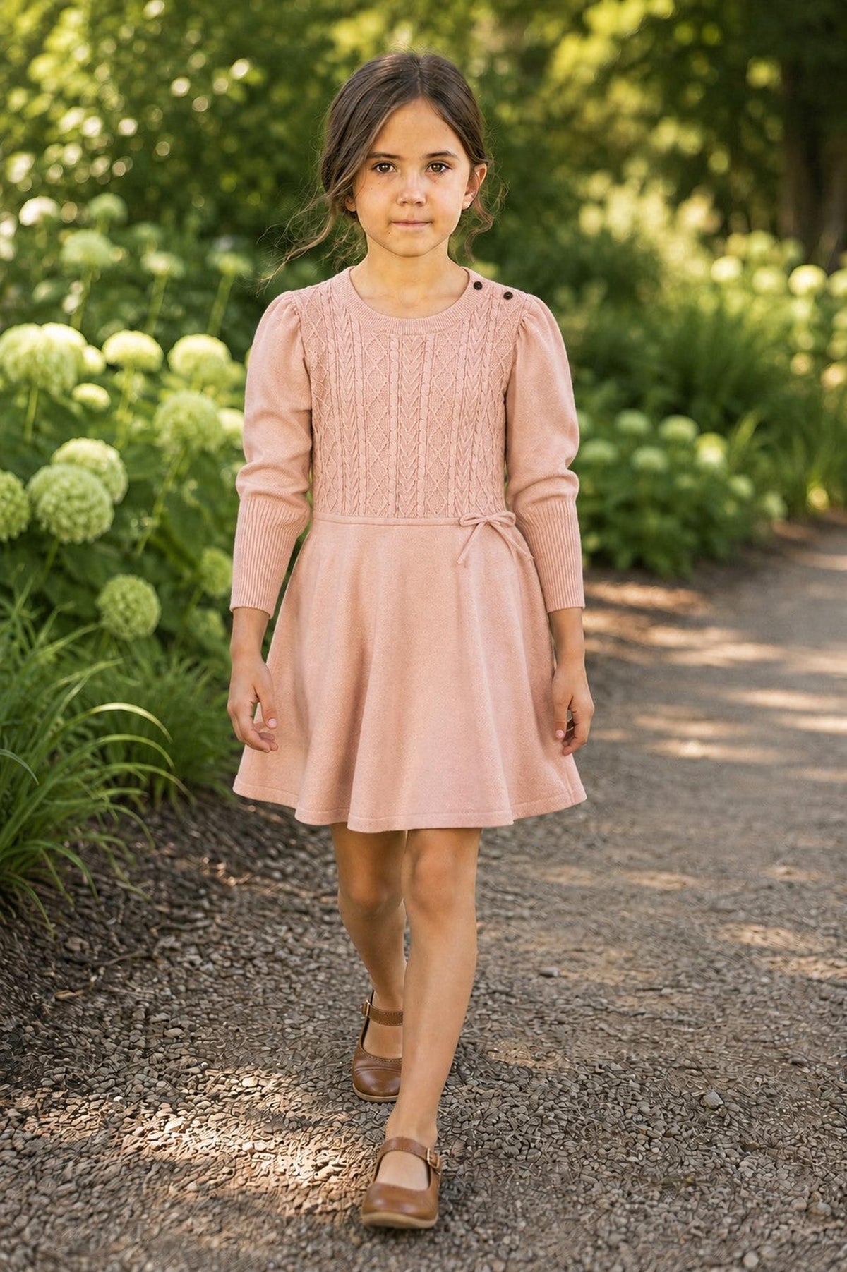 A young girl walks confidently along a garden path, wearing a blush organic cotton sweater dress with cable knit detailing and button accents on the shoulders, complemented by brown shoes.