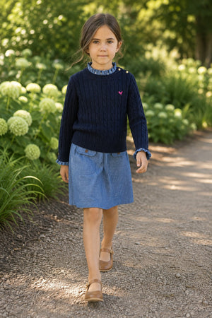 A young girl walks along a path wearing a navy cable knit sweater and a denim skirt. The setting features lush greenery and blooming flowers, highlighting a classic, stylish look.