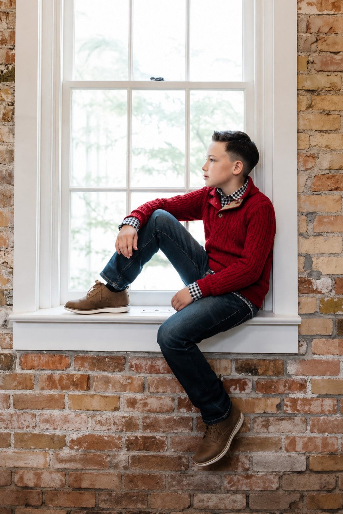 A boy sits on a windowsill, wearing a red organic cotton mock neck cable sweater and dark jeans.