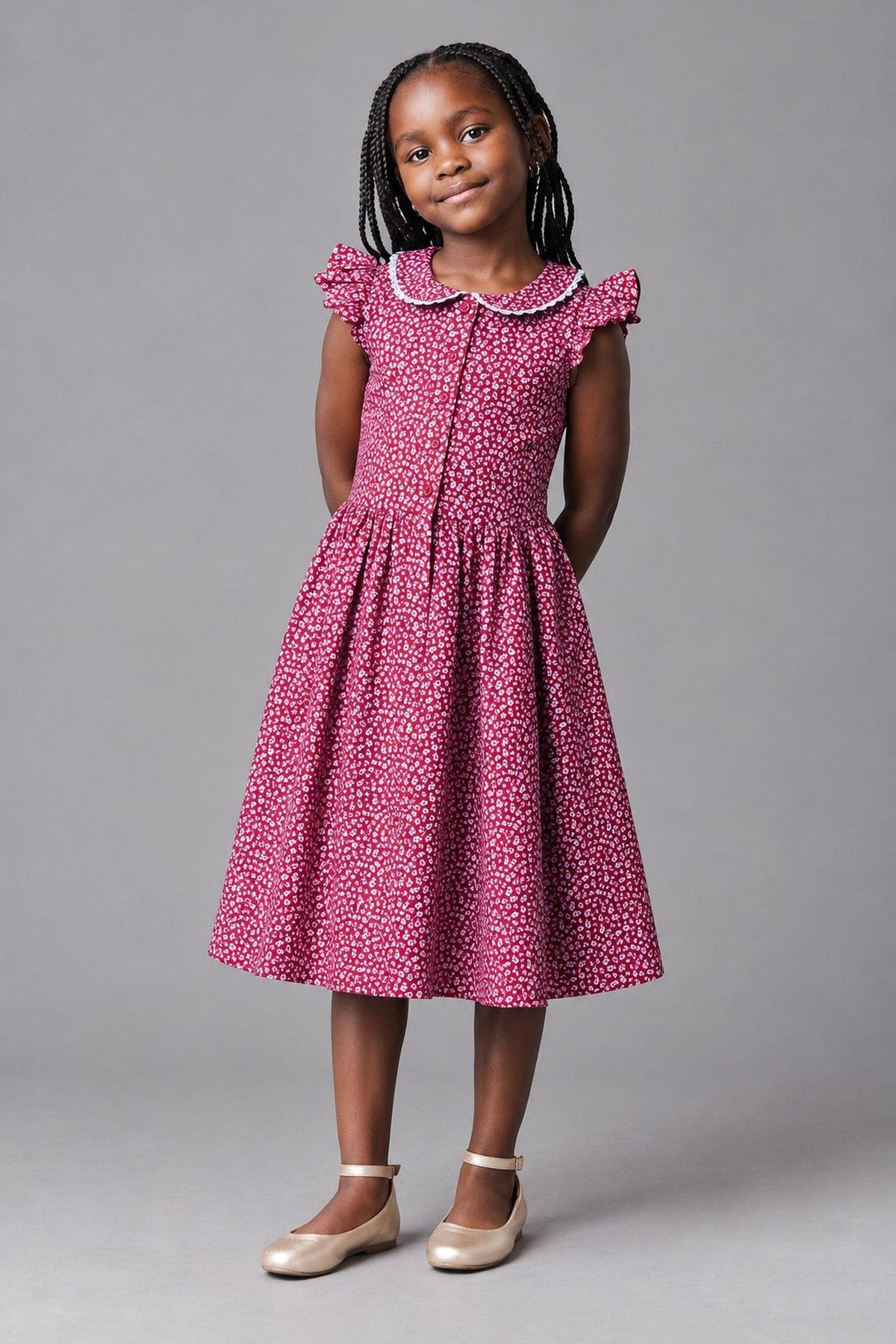 A young girl stands confidently in a wine-colored linen dress adorned with a delicate floral pattern. The dress features a Peter Pan collar, flutter sleeves, and a button front closure.