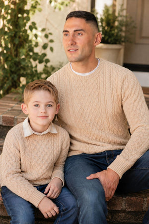 A father and son sit together on stone steps, both wearing matching organic cotton fisherman sweaters in a natural beige color, featuring an intricate cable knit design.