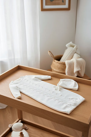 A soft white organic cotton sweater gown and bonnet set is displayed on a wooden surface, surrounded by neutral decor.