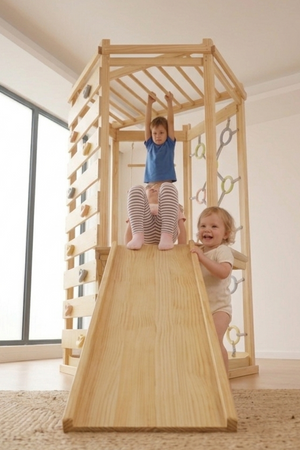 Two children play on a wooden indoor climbing gym featuring a slide and climbing elements. The structure is made from natural wood, set in a bright, spacious room.