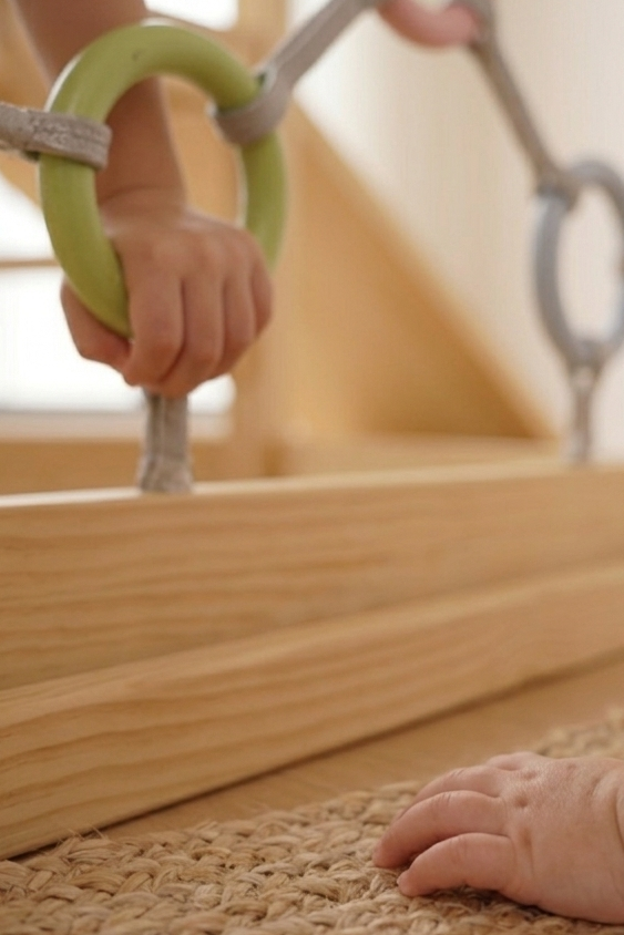 A childs hand grips a green ring on a wooden climbing gym, showcasing the natural wood finish and safe play features. The textured mat beneath adds an element of comfort.