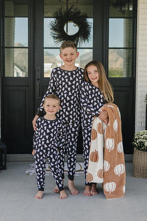 Three children wearing matching black pajamas with white ghost prints stand in front of a door, one holding a pumpkin.