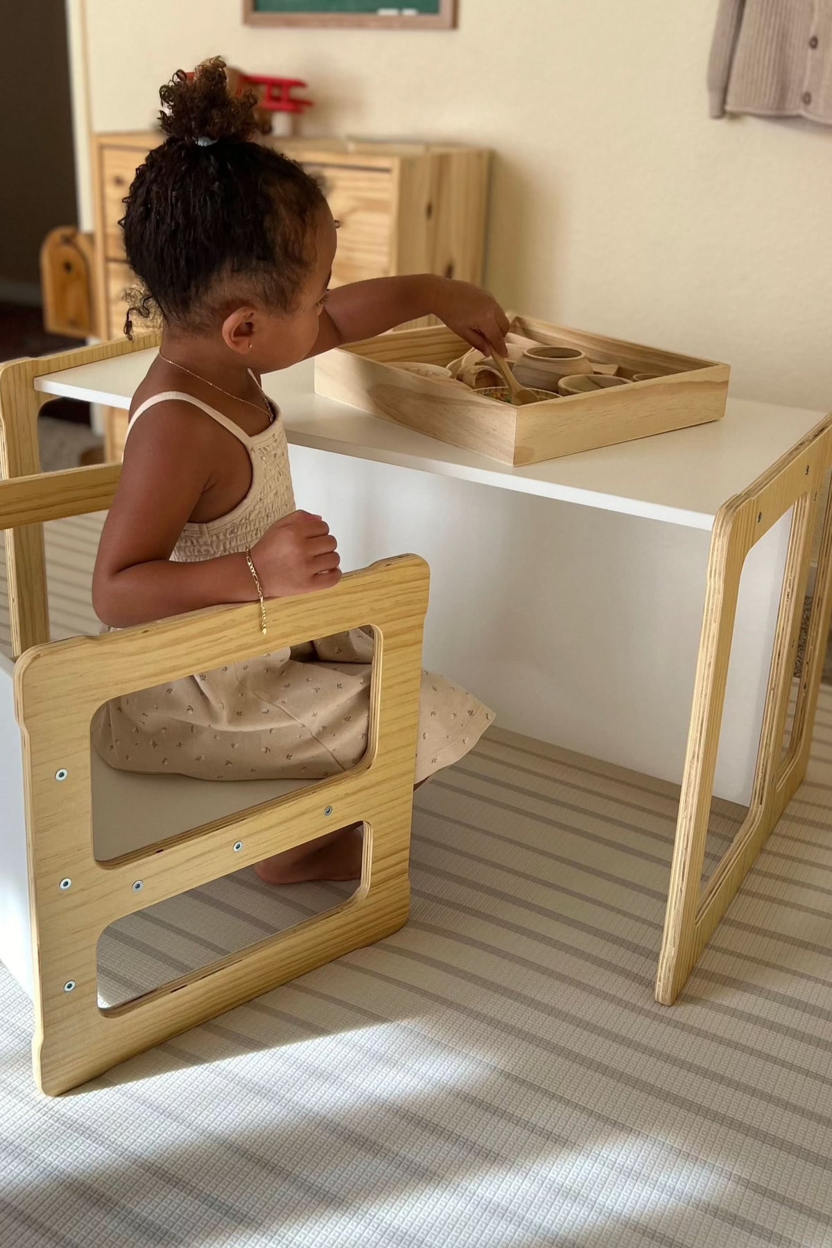 A young child sits at a modern, convertible wooden table with a white surface and natural wood accents, engaged in play with a tray of wooden toys.