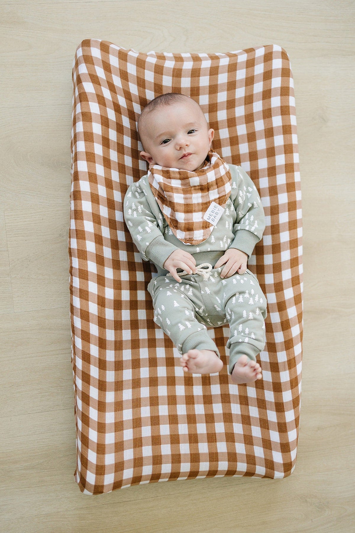 Baby lying on a brown and white gingham muslin changing pad cover, wearing green patterned outfit and matching bib.