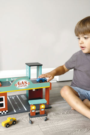 Child playing with a colorful wooden gas station toy set, featuring cars and pumps on a wooden floor.