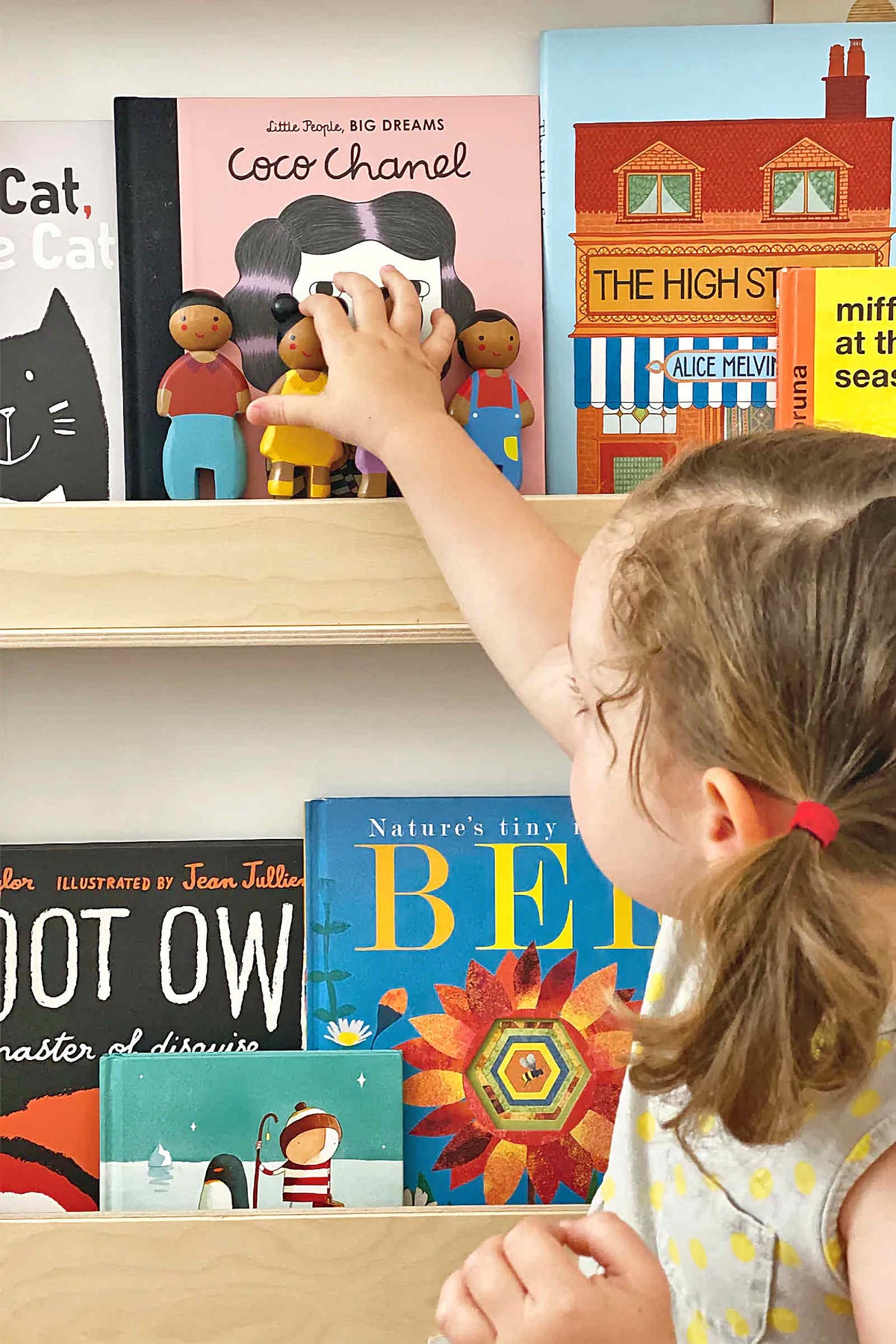 A child reaches for colorful wooden dolls on a shelf, surrounded by various childrens books in a bright setting.
