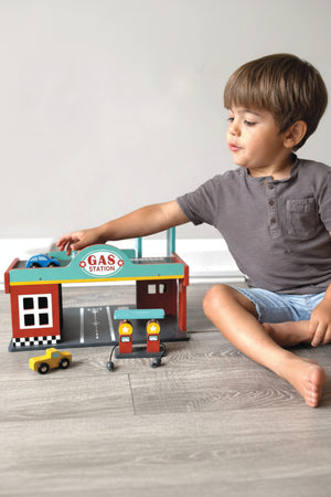 A child plays with a wooden gas station toy set, featuring colorful cars and pumps on a wooden floor.