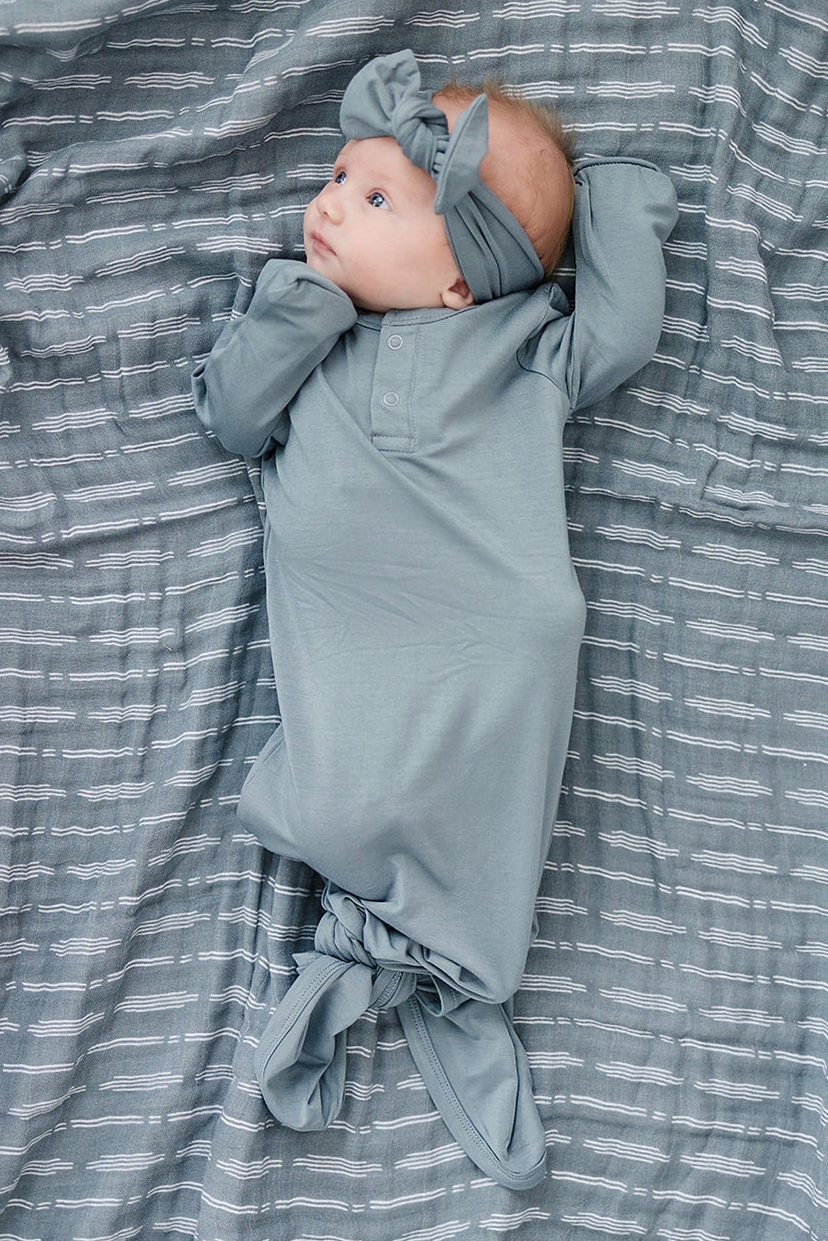Baby wearing a dusty blue head wrap and gown, lying on a matching patterned fabric.