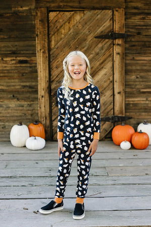 Child wearing Mebie Baby Halloween ghost print pajama set, standing on wooden porch with pumpkins in the background.