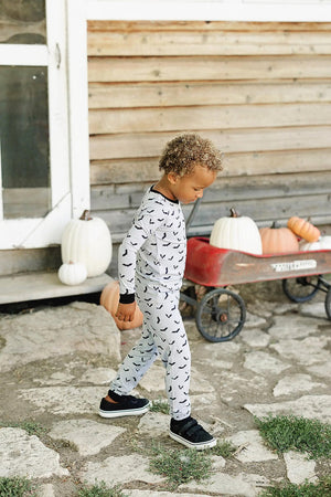 A child wearing bat-patterned pajamas walks on a stone path beside a red wagon with pumpkins.