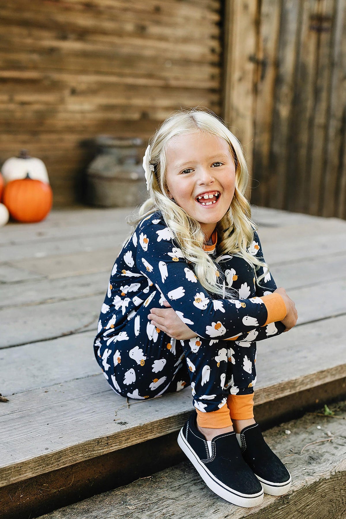 A child wearing a navy pajama set with white ghost prints sits on wooden steps, smiling.