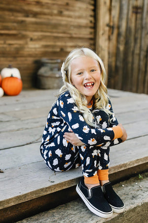 A child wearing a navy pajama set with white ghost prints sits on wooden steps, smiling.