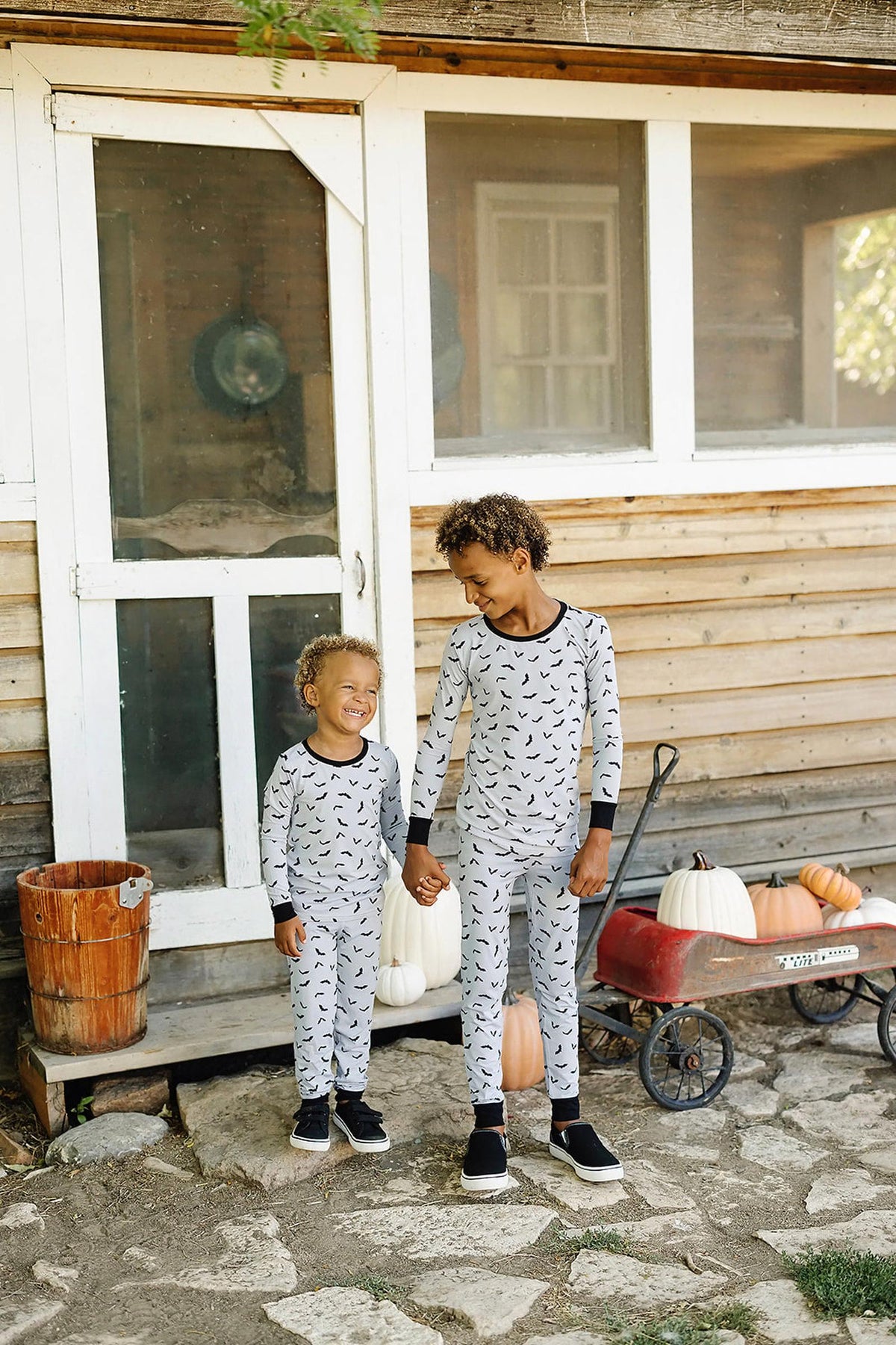 Two children wearing matching gray pajamas with a bat print stand outside a rustic house, holding hands and smiling.