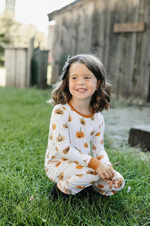 A child wearing a white pajama set with orange pumpkin prints sits on grass, smiling outdoors.