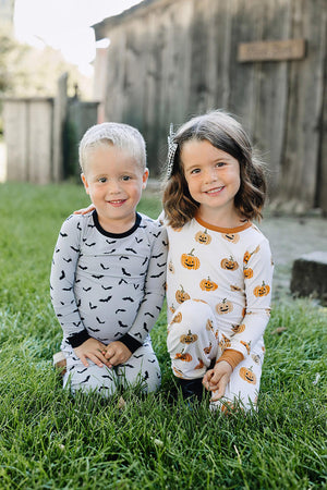 Two children kneel on grass, wearing Halloween-themed pajamas with bat and pumpkin prints, smiling outdoors.