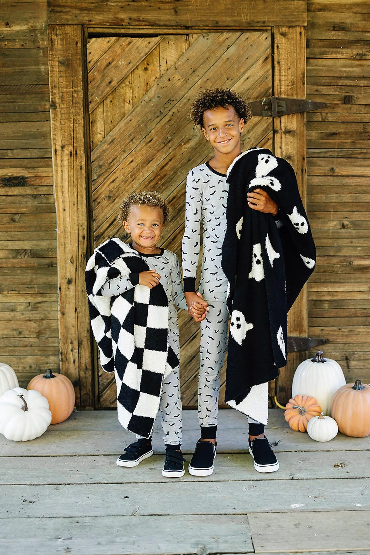Two children in bat-print pajamas hold Halloween-themed blankets, standing on a wooden porch with pumpkins.