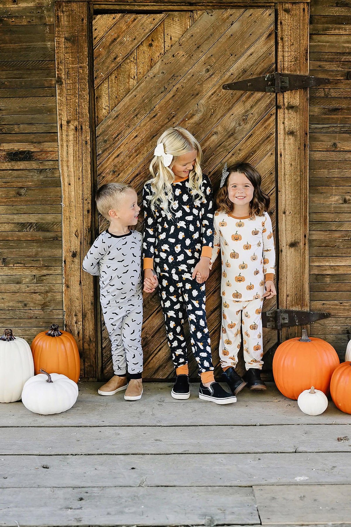 Three children wearing Halloween-themed pajamas stand in front of a rustic wooden door with pumpkins around them.