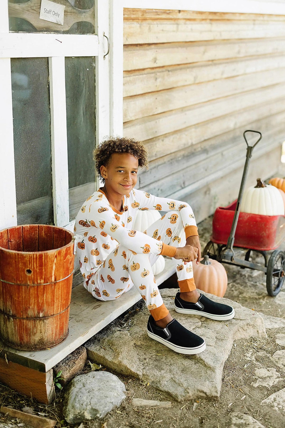 A child in pumpkin-patterned pajamas sits by a wooden wall, next to a red wagon and pumpkins.