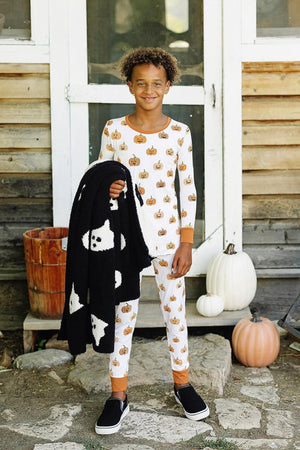 A child wearing a pumpkin-print pajama set holds a black blanket with ghost designs, standing by pumpkins.