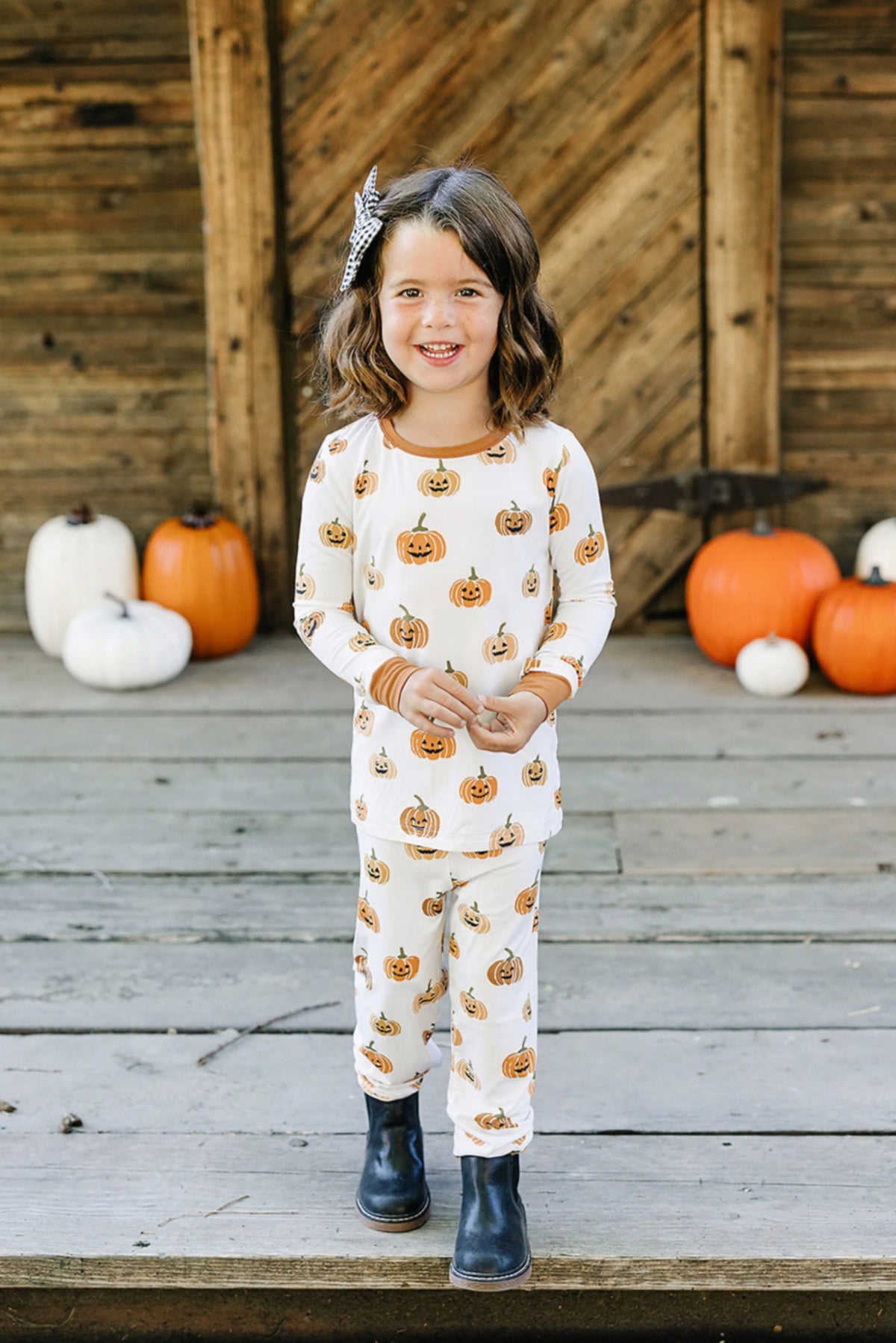 A child wearing pumpkin-patterned pajamas stands on a wooden porch with pumpkins in the background, smiling warmly.