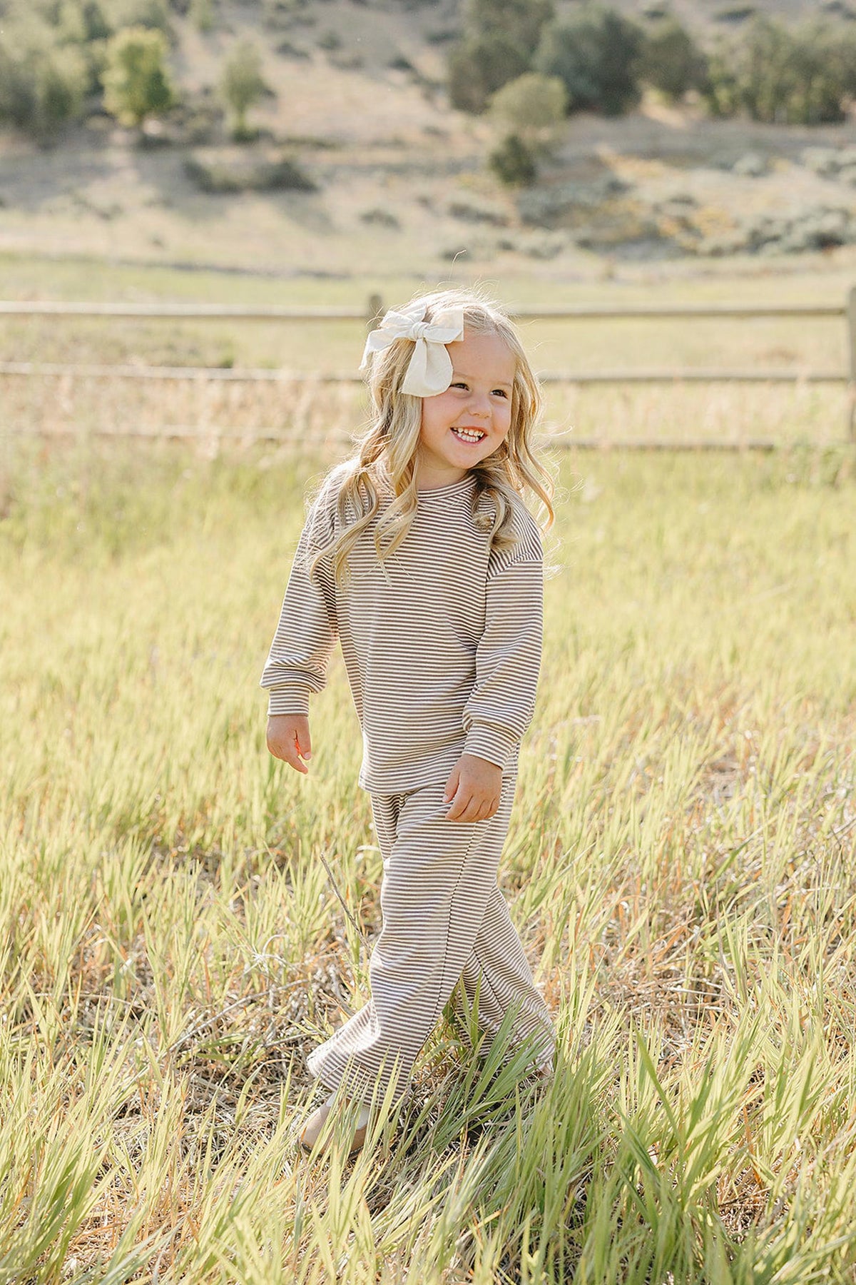 A smiling child wears a striped loungewear cotton set, featuring a long-sleeve top and pants, while standing in a grassy field with a rustic wooden fence in the background.